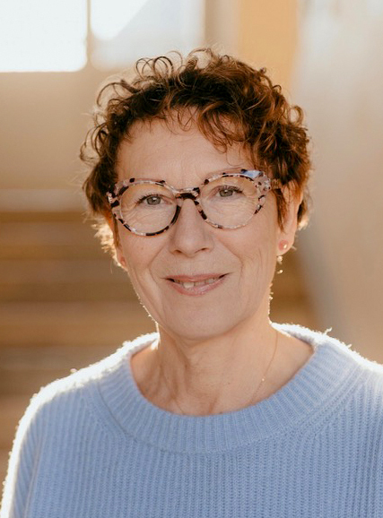 Portrait d'Isabelle Fournier, souriante, avec des lunettes à motifs, vêtue d'un pull bleu clair, reflétant la sérénité et le professionnalisme en tant qu'accompagnatrice bien-être
